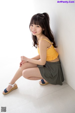 A woman in a yellow top and green skirt leaning against a wall.